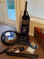 Photo showing Hoover Wind Tunnel upright vacuum in dark blue with clear dust container, flexible black hose, several attachments, and vacuum bags laid out around it.