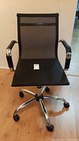 Front view of the office chair showing the mesh backrest, black cushion seat, chrome armrests with padding, and base with wheels.
