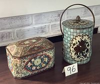 Photo showing two vintage metal tins on a wooden surface with a white brick-style background. One is cylindrical with a handle and floral/geometric pattern, the other is a rectangular tin with an octagonal top and a colorful floral design.