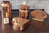 Four copper kitchen items arranged on dark surface against a decorative white brick wall, including a tall milk can, a lidded canister, a heart-shaped mold, and a napkin holder.
