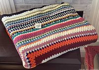 Photo of folded large multicolored crocheted afghan blanket with striped granny square pattern in various colors including white, green, pink, orange, red, brown, purple, and yellow.