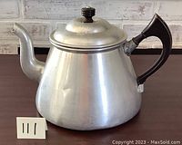 Front and side view of the large vintage aluminum teapot showing its shape, black handle and lid knob.