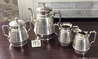 Full tea service set includes teapot, sugar bowl, creamer, and additional pitcher, displayed on a dark brown surface with a stone wall background.