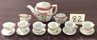Front view of entire miniature tea set displayed with 6 cups, 6 saucers, teapot, creamer, and sugar bowl with lid, showing detailed red and green decorative pattern.
