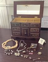 Wooden jewelry box open showing compartments, mirror inside lid, and assorted costume jewelry pieces laid out in front.
