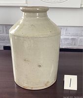 Front and side views of an old beige stoneware crock showing cylindrical body, narrow neck, and glazed surface with visible crazing and chip near base.