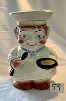 Front view of the vintage ceramic chef cookie jar, showing the face, spoon, skillet with yellow detail, and chef's hat lint lid on top.