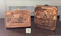 Two copper molds displaying embossed house designs side by side on a white bench.
