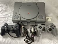 Top down view of Sony PlayStation console with closed lid, two wired controllers (black and gray) and cables visible.