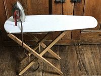 Wooden child-sized ironing board with white fabric cover and toy iron placed on top in front of wood door.