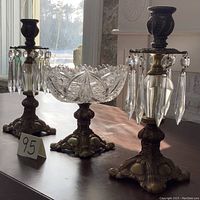 Photo showing two brass candlesticks with hanging glass prisms flanking a central crystal compote bowl on a brass base, all displayed in sunlight highlighting the prisms and crystal cuts.