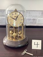 Full view of vintage brass domed clock under glass dome with winding key beside it, showing front clock face and base.