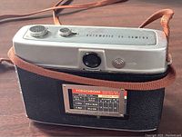 View of front and top of Kodak Automatic 35 camera sitting on original brown leather case with strap, showing markings and lens.