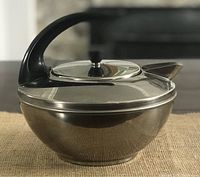 Side view of the vintage Revere Ware stainless steel kettle showing the rounded bowl-like body, lid with black knob, and uniquely shaped spout.