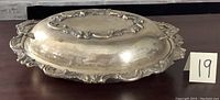 Oval silverplate serving dish with cover, showing side and top view with ornate decorative edges and central motifs. Surface tarnished and dull, needing polishing.