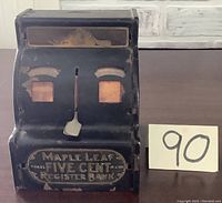 Front and side view of vintage tin bank showing wear and Maple Leaf Five Cent Register branding