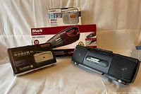 Photo showing three radios and Shark handheld vacuum placed on fabric surface; boxes of Coby CX-39 and Shark vacuum visible, Cougar and Gold Yip radios visible; confirms presence and condition of items.