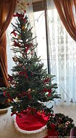 Full view of assembled 6ft artificial Christmas tree decorated with red ribbon bows, gold and vintage-style glass ornaments, draped with garland, and lit with small clear lights. Red tree skirt at the base.