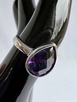 Close-up image of silver ring with large pear-cut purple amethyst gemstone set in bezel setting, displayed on black hand model showing ring shape and gemstone color.