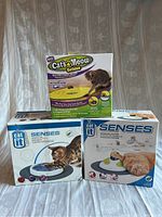Three boxed cat sensory toys: Cat's Meow Deluxe motorized wand and two Catit Senses interactive toys, on white patterned surface.
