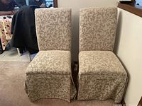 Front view of two beige upholstered dining chairs with floral damask pattern and skirted base, placed side by side.