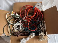 Assorted electrical cords coiled and piled inside a cardboard box showing different colors and lengths.