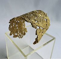 Gold-tone cuff bracelet displayed on an acrylic stand showing lace floral and leaf pattern detail from one angle.
