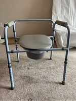 Front view of folding deluxe adjustable commode toilet showing padded armrests, seat, and removable waste bucket underneath.