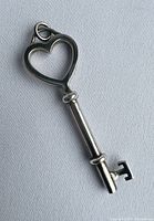 Close-up front view of silver key pendant with heart-shaped bow, showing smooth surface and loop for chain attachment.
