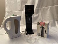 All four items displayed together on white backdrop: black SodaStream maker, white electric kettle, clear glass teapot, boxed Bialetti coffee maker.