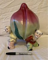 Front view of porcelain statue with three boys carrying large gradient pink and green peach, sharpie pen included for scale.