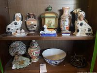 Full view of shelf with porcelain Asian figurines, vases, trinket dishes, candle holder, decorative plates, serving bowl.