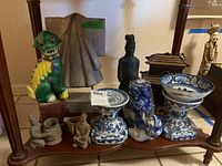 Wide shot showing the arrangement of all items including Foo dogs, miniature figurines, candle holders, cloak, wood house, and bowl on a wooden shelf.