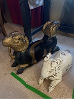 Photo showing two wooden goat statues, one black with gold accents and a larger size, and one smaller white with black accents, both sitting on beige carpet.