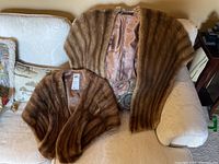 Both mink shawls displayed side by side on white couch. One larger shawl with scalloped fur pattern, other smaller with pockets visible.