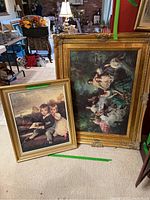 Photo showing both framed paintings placed on the floor, the larger one with an ornate gold frame and damage visible at corners.