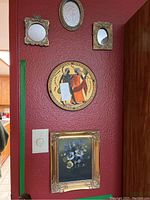 Overall view of the wall installation featuring one framed floral painting, one decorative round classical style plate, and three small ornate framed mirrors on a dark red wall