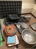 Photo of various muffin tins, round baking pans, heart-shaped cake pan, rectangular baking pans, metal colander, and fabric trivets arranged on a floor.