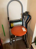 Photo showing chrome stool with round orange seat, folding step stool behind, and canes standing together
