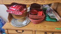 Wooden sideboard top with folded Christmas-themed tablecloths, clear glass serving platter, woven baskets with red cups, and some folded green plasticware items.
