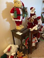 View of various Christmas figures including a large plush bear dressed as Santa Claus holding a Christmas tree branch and sack, Mrs. Santa figurines, and small decorations arranged on shelves and tables.