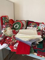 Photo displaying assorted Christmas tablecloths, napkins with poinsettia patterns, Santa figurines, and festive pillows on a table.