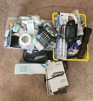 Top-down photo of two containers filled with various small items including floral tape, fastener dots, velcro, packaging tape, and small boxed items.