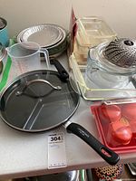 Overview of the lot showing skillet with glass lid, Pyrex glass casserole pans, metal steamer basket, aluminum pie plates, plastic measuring cup, and tomato-shaped plastic containers.