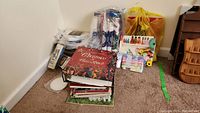 Photo showing assorted cross stitch items stacked on carpet including books, fabric bundles, and plastic bags of supplies.