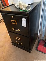 Side angled view of black two drawer filing cabinet with brass handles and label holders, showing wear marks and top surface with miscellaneous items (not included).