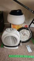 Sunbeam humidifier, desk fan heater, and wall clock grouped together on carpeted floor with a white humidifier on top, black wall clock in middle, and white desk fan heater in front.