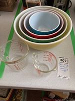 Photo showing set of six nested Pyrex mixing bowls in different colors and two glass measuring cups with red measurement markings sitting on a table.