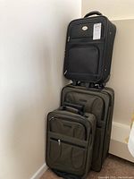 Three travel bags stacked: two olive green carryon suitcases and one black carryon bag on top.
