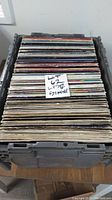 Stack of vinyl records in sleeves stored upright in a plastic crate with a handwritten sign 'LOT 67 LP 12 Single'.